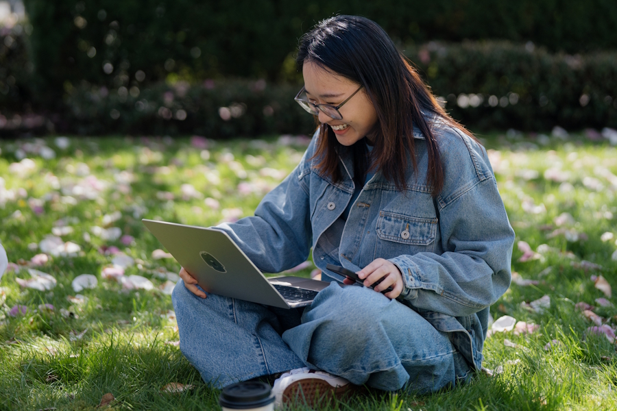A young woman sitting on a flower-strewn lawn smiles at what she sees on her laptop screen